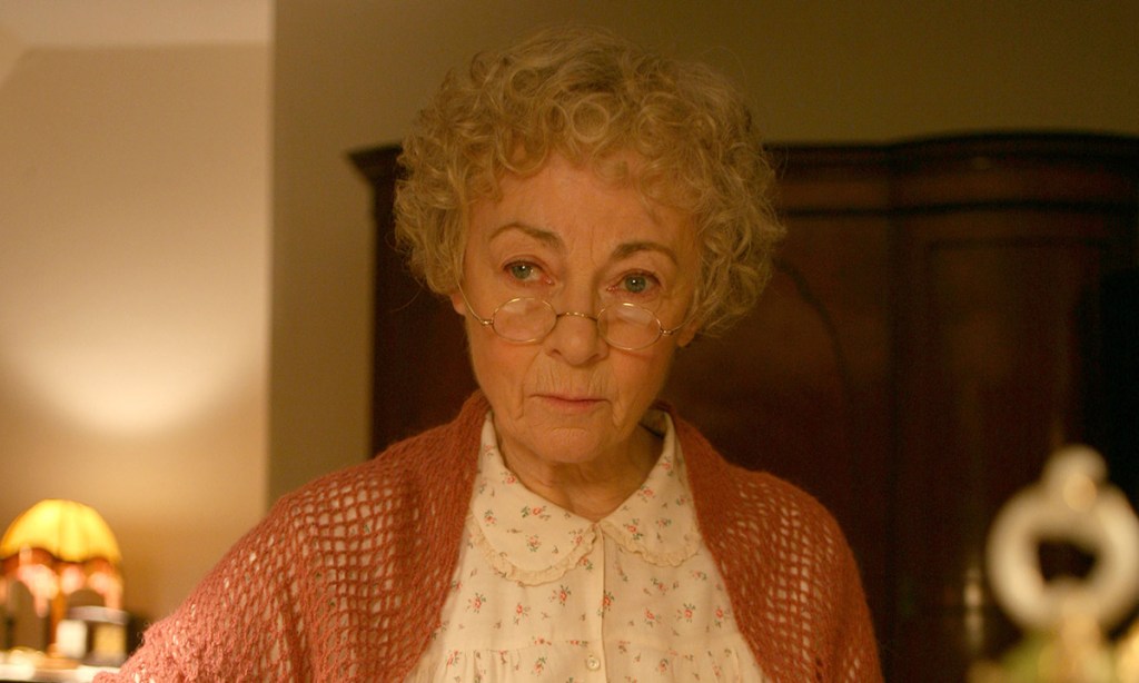 Actress Geraldine McEwan in a still from the Masterpiece Mystery series Miss Marple. She has tightly curled white hair, gold-rimmed glasses, a floral old-fashioned top, and a rose-colored shawl.