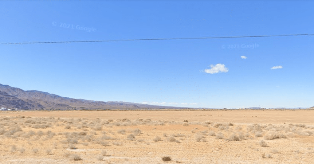 A screenshot of the Mojave Desert as seen by Google Earth outside Newberry Springs, California. There is a wide blue sky with three small puffy clouds, all above a wide tan desert with some assorted small scrub. Along the horizon is the dark gray-brown rise of a mountain to the left. A powerline cuts across the upper portion of the picture.