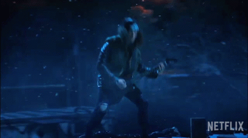 A gif of Eddie Munson as he shreds guitar in the Upside Down during Stranger Things Season 4. The background is dark and bits of ash swirl around him, but he is a head-banging metal hero despite it all.